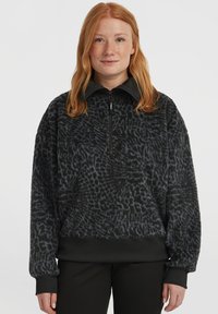 Vrouw met rood haar, gekleed in een zwarte structuur fleece pullover met een kwart rits en geribde manchetten en een zoom, staand tegen een witte achtergrond.