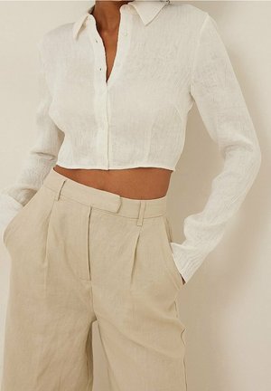 Femme portant une chemise courte blanche texturée à boutons avec manches longues et un pantalon plissé beige taille haute, mains dans les poches.