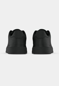 Fekete, alacsony szárú sportcipő pár, hátulról fényképezve, texturált talppal és sima matt felülettel, fehér háttér előtt.