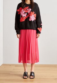 Svart stickad tröja med blommig broderi i rosa och rött; tillsammans med en plisserad rosa kjol och svarta slidesandaler.