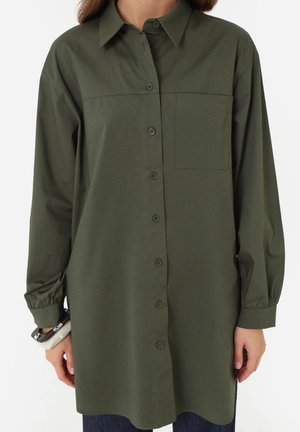 Kvinde iført en oversized olivengrøn skjorte med brystlomme og lange ærmer, der holder armbånd på venstre håndled.