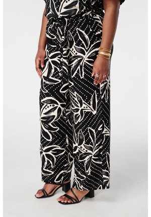Femme portant un pantalon noir à jambes larges avec un grand imprimé floral blanc et des sandales à talons à brides noires, montrant le bas du torse et les jambes.