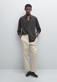 Dark gray button-up shirt with rolled sleeves, paired with beige pleated trousers and black suede shoes. Model standing with hands in pockets.