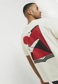 Cremefarbenes T-Shirt mit einem großen rot-schwarzen abstrakten Grafikdesign auf der Rückseite, mit rundem Ausschnitt und kurzen Ärmeln.