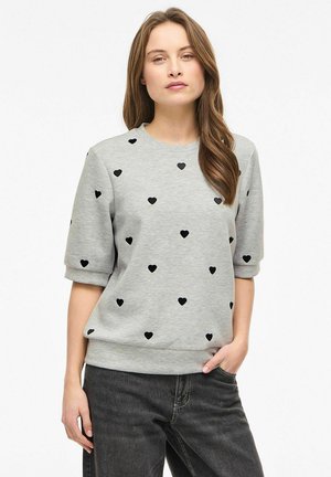 Ung kvinde iført en grå sweatshirt med korte ærmer og sorte hjerteprint samt mørkegrå jeans, stående med den ene hånd i lommen.