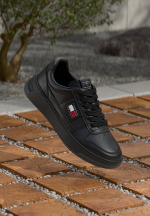 Czarny sneaker Tommy Jeans z sznurowadłami, pokazany w powietrzu nad drewnianymi płytkami i żwirem, z rozmytym drzewem i szarym tłem.