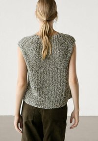 Top senza maniche grigio a maglia con vestibilità ampia, motivo testurizzato e orlo arrotondato, abbinato a pantaloni scuri. Vista posteriore, sfondo minimalista.