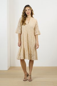 Beige spetsklänning med v-ringning, utsväva ärmar och utsvängd kjol, prydd med blommönster. Matchat med gröna sandaler med remmar och hög klack.