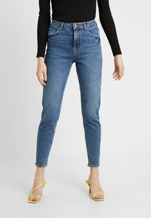 Slim fit blå jeans med høj talje båret med sort langærmet top og gule åbne hælede sandaler på hvid baggrund.