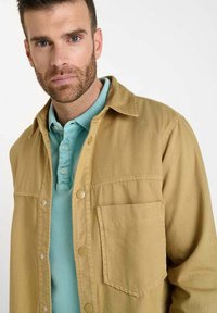Veste boutonnée beige avec une poche poitrine superposée à une chemise polo bleu clair. Le tissu semble lisse, avec des couleurs contrastantes et un design simple.