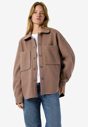 Femme portant une veste marron ample à boutons avec de grandes poches devant, par-dessus une chemise blanche et un jean bleu, debout devant un fond uni.