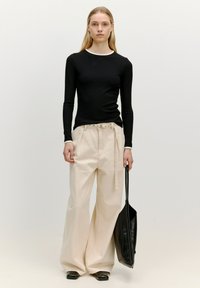 Zwarte geribbelde lange mouw top met een witte halslijn, gecombineerd met lichtbeige wijde broek. Houdt een zwarte tote bag vast.