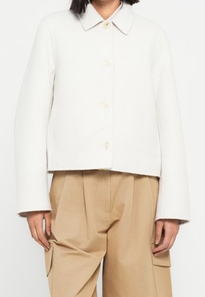 Personne portant une veste blanche boutonnée avec un col et un pantalon beige à jambes larges avec de grandes poches latérales.