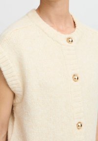 Cárdigan color crema de manga corta, confeccionado con un material de punto suave. Presenta botones dorados y un detalle de cuello acanalado.