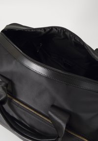 Fekete utazótáska cipzárral a tetején. Texturált külső, sima belső arany cipzáras zsebbel. Két tartós fogantyú az oldalán.