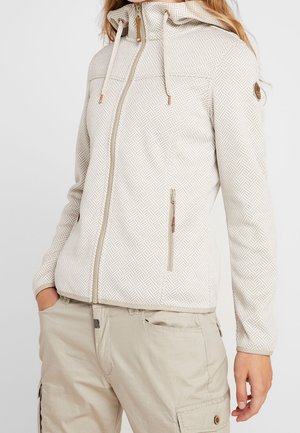 Woman wearing a light beige patterned zip-up jacket with hood and beige cargo pants, with hands in pockets, against a plain background.