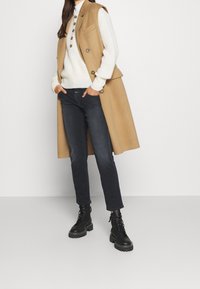 Gilet lungo beige con bottoni, maglione bianco a maglia, jeans scuri e stivali neri con lacci e suole spesse. Design minimalista, colori neutri.