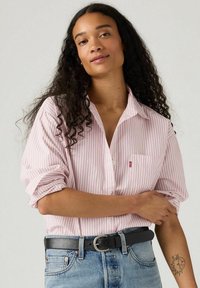 Camicia a righe rosa e bianca con maniche arrotolate, tasca sul petto e una piccola etichetta con logo, abbinata a jeans di denim a vita alta.