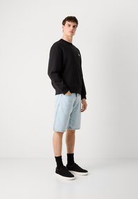 Zwart sweatshirt met ronde hals, lichtblauwe denim shorts en zwarte sneakers. Model staat met handen in de zakken tegen een witte achtergrond.