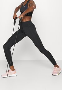 Leggings neri per il fitness con una vita alta, abbinati a un reggiseno sportivo nero e scarpe da ginnastica rosa chiaro, mentre si utilizzano fasce di resistenza.