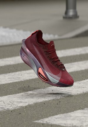 Chaussure de sport marron avec des accents blancs et roses flottant au-dessus d'un passage pour piétons sur une route en asphalte.