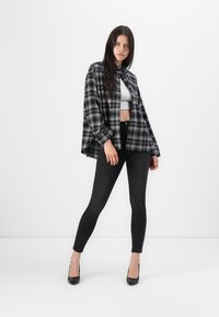 Camicia oversized a quadri bianco e nero con maniche lunghe, indossata sopra un top corto bianco e abbinata a jeans skinny neri e tacchi alti.