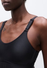 Κοντινό πλάνο ενός ατόμου που φοράει μαύρο αθλητικό σουτιέν με ρυθμιζόμενους ιμάντες Under Armour και ένα μικρό τατουάζ στον ώμο εμφανές.