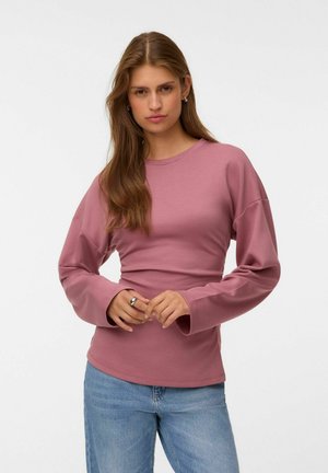 Jeune femme aux longs cheveux bruns portant un haut à manches longues rose et un jean bleu, debout devant un fond clair uni.