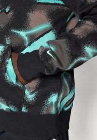 Sudadera negra con diseño salpicado en tonos verde azulado y marrón. Cuenta con un bolsillo tipo canguro, puños acanalados y un pequeño logotipo blanco de Nike en la manga.