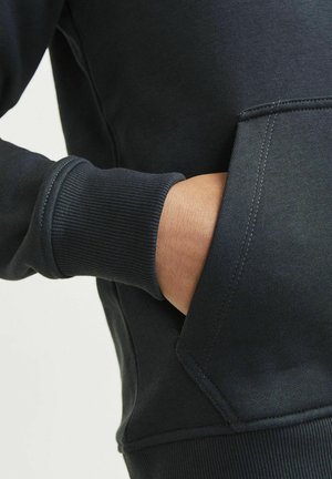 Felpa blu scuro con polsini a coste e tasca frontale. Il tessuto è liscio con cuciture visibili lungo i bordi e le cuciture.