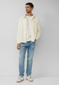 Hellbeiges, strukturiertes Hemd mit Knöpfen über einem weißen T-Shirt, kombiniert mit blauen Jeans und beigen Sneakers, stehend vor einem neutralen Hintergrund.