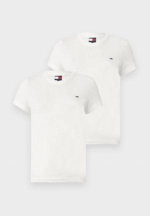 Νεαρή γυναίκα με σγουρά μαλλιά φοράει εφαρμοστό λευκό μπλουζάκι, άνετο ανοιχτό μπλε τζιν και λευκά αθλητικά παπούτσια, στέκεται μπροστά σε μια απλή φόντο.