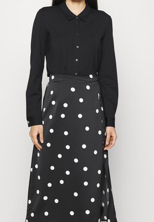Femme portant une chemise noire à manches longues boutonnée, rentrée dans une jupe noire à pois blancs avec une fente sur le côté.