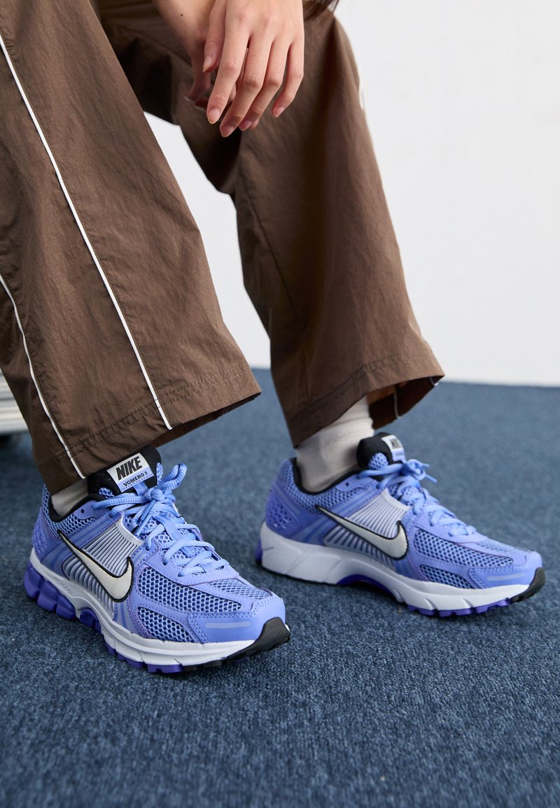 Sapatilhas de corrida Nike em azul claro e malha cinza, com um superior texturizado, realces em preto e solas roxas, usadas com calças castanhas.