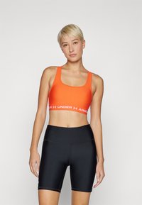 Oransje sports-bh med brede stropper og hvit logo-bånd, paret med høy midje svarte sykkelshorts. Jevn tekstur og tettsittende design.
