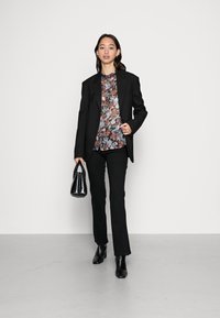 Blazer preto ajustado sobre uma blusa de padrão floral em azul, vermelho e branco. Calças pretas justas e botins pretos de salto, segurando uma mala preta.