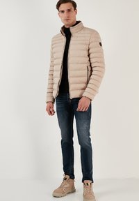 Lys beige pufferjakke med vandret quiltning, høj krave og lynlås foran. Parret med mørke jeans og beige høj-top sneakers.