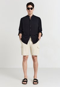 Schwarzes Langarmshirt mit Knopfleiste, beigefarbene Cargo-Shorts und schwarze Sandalen. Einfaches Design mit lockerer Passform und niedrigem Kragen.