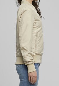 Kvinna klädd i en beige bomberjacka och blå jeans, stående i profil mot en enkel ljusgrå bakgrund.