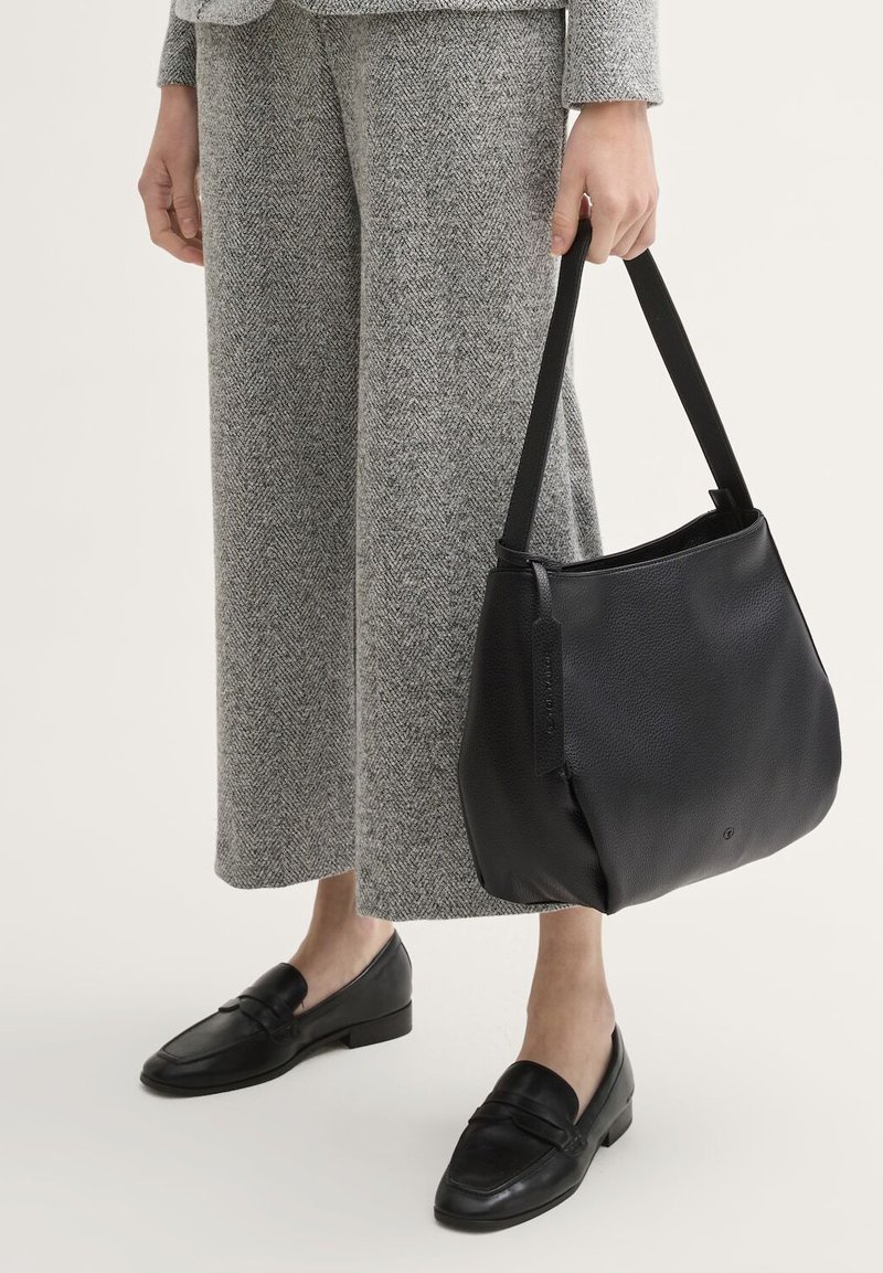 Sac en cuir noir avec une finition douce et texturée et une seule sangle noire. Porté avec un pantalon en chevrons gris et des mocassins noirs.