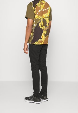 Muž v košili s krátkým rukávem s žlutým barokním vzorem, černých kalhotách a černých botách Versace stojí proti jednoduchému pozadí.