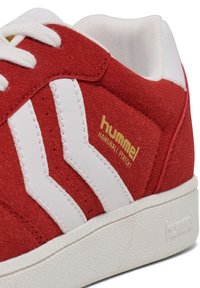 Red suede athletic shoe with white leather accents, featuring three white stripes, gold branding, and a textured white sole.
