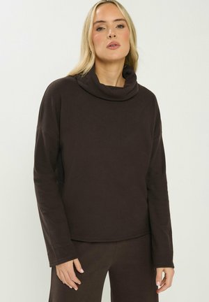 Frau trägt ein dunkelbraunes Langarm-Oberteil mit Rollkragen und passende dunkelbraune Hose vor hellem, einfarbigem Hintergrund.