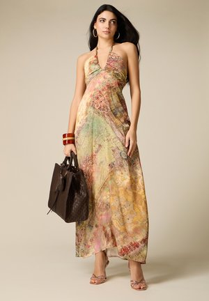 Donna che indossa un abito lungo senza maniche multicolore con collo halter, tiene una grande borsa intrecciata marrone, accessoriata con braccialetti e orecchini a cerchio.