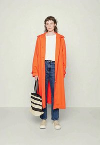 Άτομο με φωτεινό πορτοκαλί παλτό, λευκή μπλούζα, μπλε τζιν, μπεζ αθλητικά πάνινα παπούτσια, κρατώντας μαύρη-άσπρη ριγέ τσάντα, στέκεται μπροστά σε απλό φόντο.