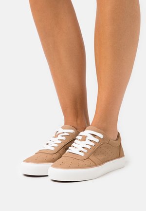 Voeten met zandkleurige suède sneakers met witte veters en dikke witte rubberen zolen op een neutrale achtergrond.