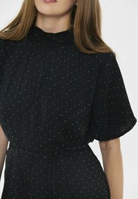 Vestido negro de manga corta, con un cuello alto. La tela es ligera con patrones de lunares blancos y un cinturón de lazo en la cintura.