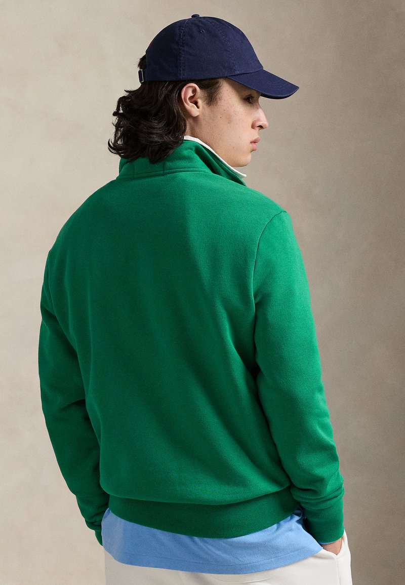 Green sweatshirt with high collar, long sleeves, and ribbed cuffs. Worn over a light blue shirt. Dark blue cap completes the look.