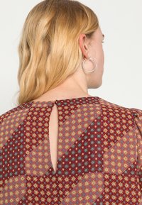 Blusa de fundo grená com padrão de pequenas flores multicoloridas, detalhe em formato de olho-chave nas costas e tecido macio e leve. Brincos circulares prateados.