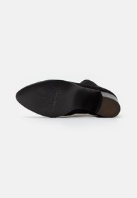Suela de zapato de ante negro con un tacón plano, que presenta una superficie texturizada y un logo en relieve. Transición suave a un acento de tacón brillante.